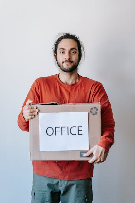 Two individuals are engaged in packing and organizing items for a home relocation or office move inside a bright room with natural light. The person on the left, with dark, curly hair and a beard, is