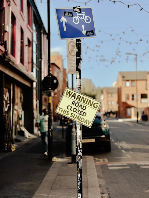 A close-up view of a rectangular road closure sign with a white background and bold black text reading 'ROAD CLOSED,' placed on a gravel surface outdoors. The sign is supported by a black plastic base