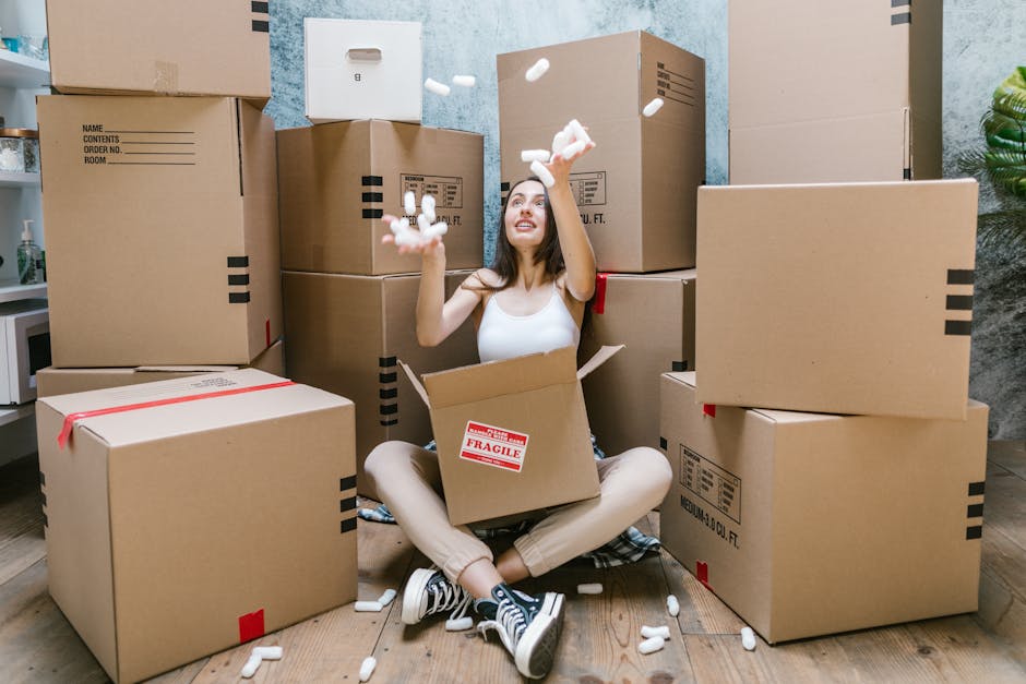 A young woman with long dark hair sits on a wooden floor inside a room surrounded by numerous unpacked cardboard boxes of various sizes, some with red 'FRAGILE' labels and others with printed shipping